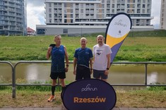 Biegacze z Klubu Biegacza PRz na tle banerów reklamowych ParkRun Rzeszów. Stoją Jan Rzucidło, Robert Jakubowski i Mariusz Węglarski i 